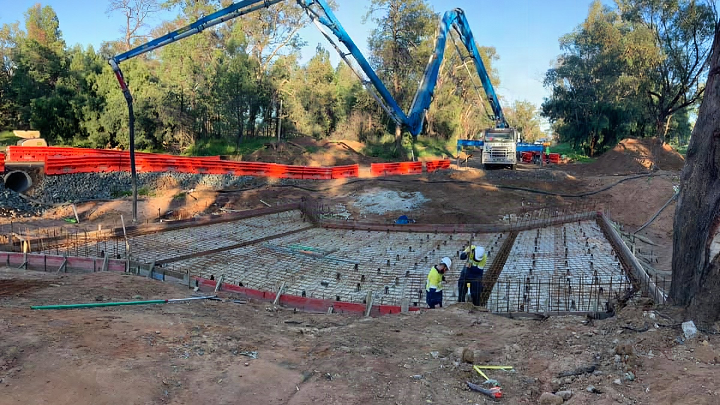 Two concrete pump booms reaching over a large formed and reinforced base cut into red soil