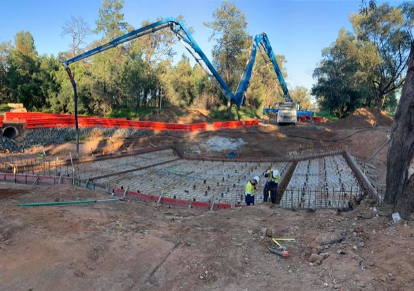 Two concrete pump booms reaching over a formed and reinforced base cut into red soil