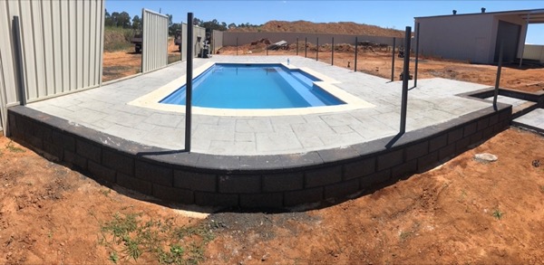 A concrete pool surround on a block retaining wall beside a shed